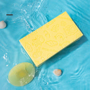 Yellow silicone baby shampoo brush with soft bristles and a bath sponge on blue background with seashells.