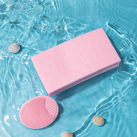 Pink silicone baby shampoo brush with soft bristles for gentle scalp cleaning and massage, shown in blue water with seashells.